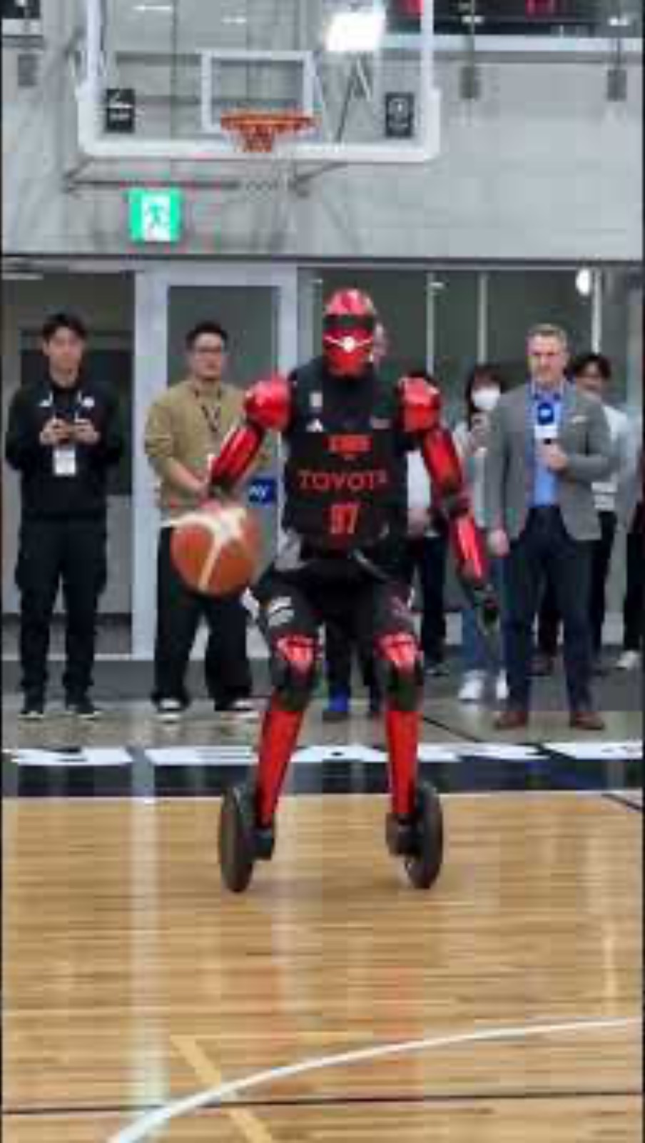 A wheeled humanoid basketball robot wearing a Toyota jersey dribbles on a hardwood court in front of onlookers